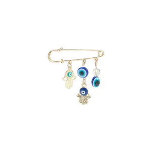 Detailed view of a gold safety pin brooch featuring blue Evil Eye beads and Hamsa hand charms.