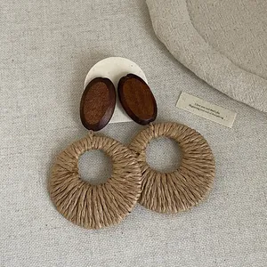 Bohemian holiday style earrings featuring dark wood studs and large woven straw hoop pendants.