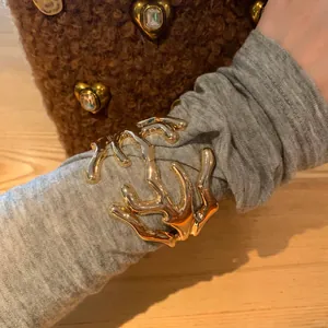 Dettaglio di un bracciale rigido dorato in metallo fuso sopra una manica grigia.