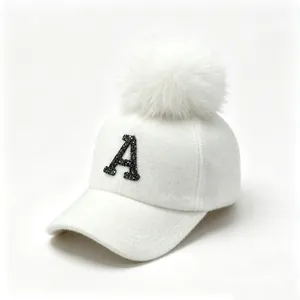 Close-up of a white felt baseball cap featuring a black rhinestone A and fur pom-pom