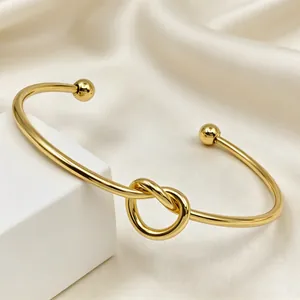 Dettaglio di un bracciale rigido aperto in acciaio inossidabile dorato con nodo centrale.