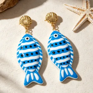 Détail des boucles d'oreilles poisson tropical avec clous coquillage doré sur sable de plage.