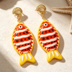 Détails des boucles d'oreilles poisson tropical avec puces coquillages dorés sur sable.