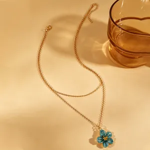 Detail shot of a layered gold necklace with a blue enamel flower pendant on a warm beige surface