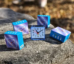 Compact blue square analog alarm clock with packaging boxes on a textured rock surface