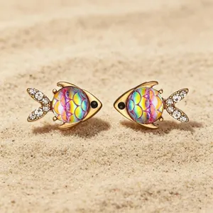 Dettaglio orecchini pesce tropicale con scaglie arcobaleno e code di cristallo sulla sabbia