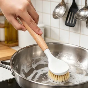 Wooden handle kitchen brush scrubbing a stainless steel pan with soap suds.