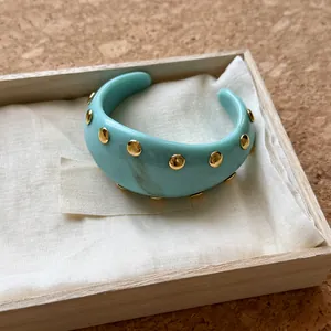 Vista detallada de brazalete ancho de acrílico jaspeado azul claro con tachuelas doradas.