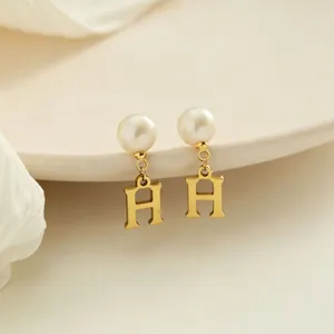 Dettaglio degli orecchini pendenti con perle e lettera H oro su sfondo crema minimal
