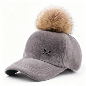 Vista lateral de gorra de béisbol gris peluda con gran pompón de piel y logotipo M plateado.