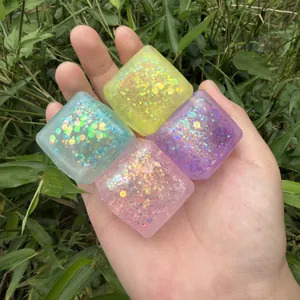 Hand holding four square glitter squeeze toys against a lush green outdoor background.