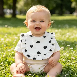 Smiling baby wearing a white ruffled gauze bib with black heart patterns in a park