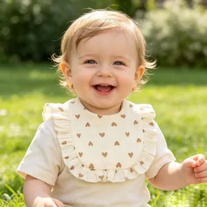 Close-up of a smiling baby wearing a ruffled cotton bib with brown heart patterns in a garden.