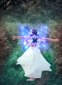 Girl wearing glowing blue LED butterfly wings and white dress in a magical forest path
