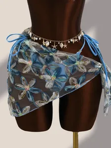 Détail de broderies florales et sequins sur paréo en mesh bleu avec chaîne de taille coquillage.