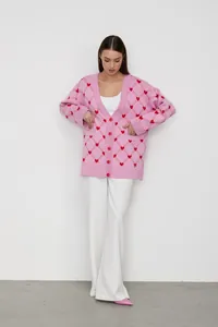 Full-length fashion view of a pink knit cardigan with red heart jacquard and white trousers.