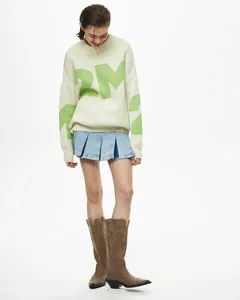 Vollansicht eines Modells in einem cremefarbenen Pullover mit grünen Buchstaben