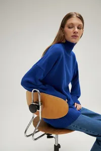 Side view of model wearing loose electric blue high-neck knit sweater sitting on wooden chair
