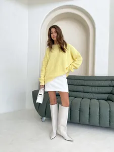 Woman wearing a loose yellow crewneck knit sweater with white mini skirt and boots in a minimalist room