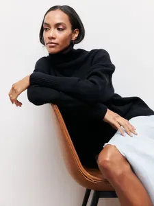 Gros plan d'un pull à col roulé noir ample porté par un mannequin dans un décor de studio sobre.