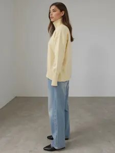 Vue de côté d'une femme portant un pull à col roulé ample jaune pâle et un jean bleu clair.
