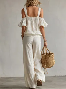 Back view of woman in white textured off-shoulder top and wide-leg trousers with straw bag.