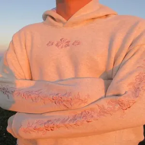 Detailed view of pink floral embroidery on sleeves and chest of a cream hoodie.