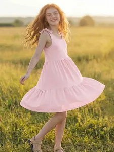 A smiling girl wears a pink sleeveless tiered dress in a sunlit field detail view.