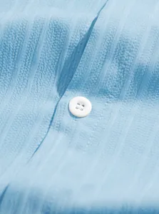 Dettaglio ravvicinato di bottone bianco e tessuto azzurro testurizzato su camicia uomo