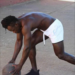 Sportler in weißen schnelltrocknenden Shorts hockt mit Basketball auf einem Außenplatz.