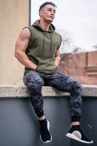 Male fitness model wearing army green sleeveless hoodie and camouflage joggers outdoors.