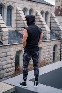 Back view of an athletic man in a black hooded gym vest and camouflage joggers outdoors.