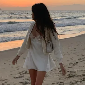 Model in witte satijnen slipdress met kant en vest op het strand bij zonsondergang