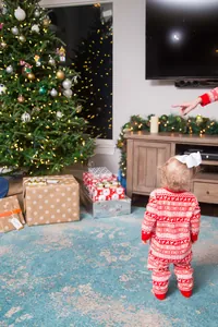 Peuter in feestelijke rood-witte kerstpyjama staat bij een versierde kerstboom in huis