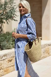 Model wearing a blue and white striped shirt dress with a straw tote in sunny outdoor setting