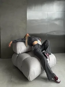Woman in dark grey tracksuit set reclining on white modular sofa in minimalist room.