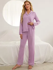 Women's long-sleeved purple pajama set with white piping in a bright bedroom setting.