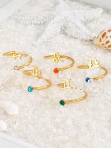 Adjustable gold-plated zodiac rings set with colorful gemstones on a white sand background