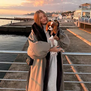 Long khaki trench coat with elegant black fur collar shown in a scenic outdoor sunset setting