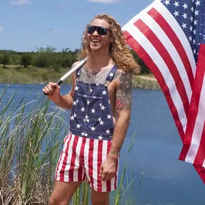 Man wearing American flag patriotic overalls holding a US flag at a sunny lakeside outdoor setting