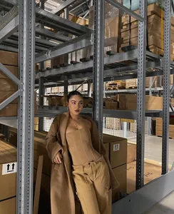 Full body view of a woman wearing a tan mesh corset top and matching coat in a warehouse.