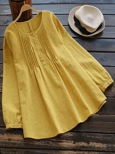 Yellow loose linen shirt with pleated details and buttoned round neck on wooden background.