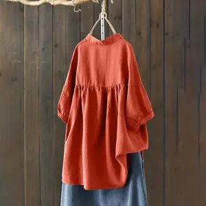 Back view of a pleated orange-red linen blouse with mandarin collar hanging on a wooden wall
