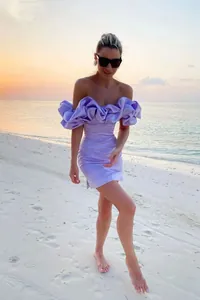 Mujer con vestido de volantes lila con hombros descubiertos en la playa al atardecer.