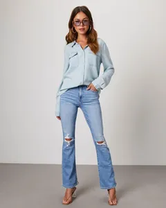 Woman in a light blue long-sleeve denim shirt and ripped jeans in a studio setting.