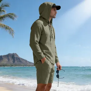 Man in olijfgroene linnen set met capuchon poseren op zonnig strand met bergen op achtergrond