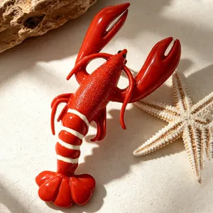 Detailed macro view of a glossy red lobster brooch resting on a sandy beach scene.