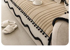 Détail d'une housse de canapé en peluche crème avec bordure ondulée à pompons noirs.