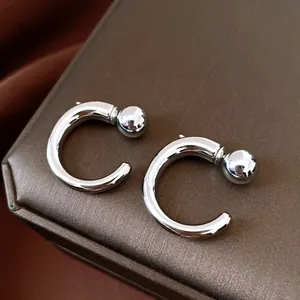 Minimalistische silberne C-förmige Creolen mit Kugel-Detail auf einer braunen Schmuckschatulle.