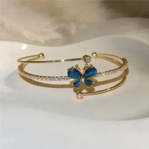 Detalle de brazalete abierto dorado con mariposa de cristal azul y circonitas brillantes.